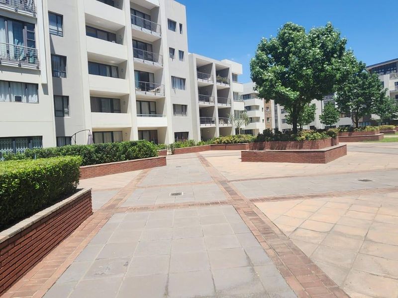 A well-maintained communal courtyard area featuring paved walkways, brick planter boxes, and landscaped hedges set against a modern multi-story townhouse complex.