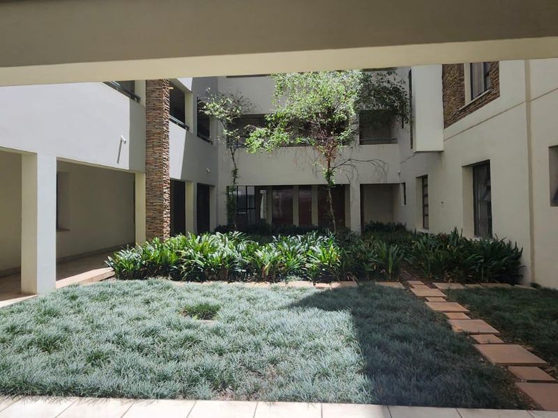A view of a well-maintained communal courtyard garden featuring stone-clad pillars, paved pathways, and lush landscaping, viewed from a covered patio area.
