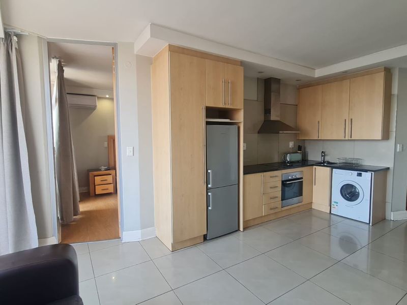 Open plan kitchen and living area featuring light wood cabinetry, integrated appliances, tiled flooring, and direct access to an adjoining room and balcony.