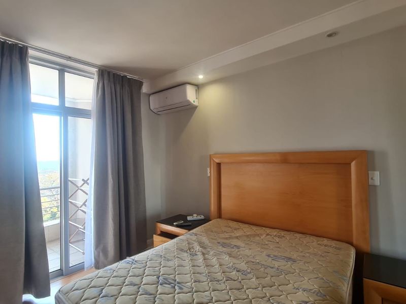 A well-lit bedroom featuring a wall-mounted air conditioning unit, recessed ceiling lighting, and a sliding glass door providing access to an outdoor balcony.