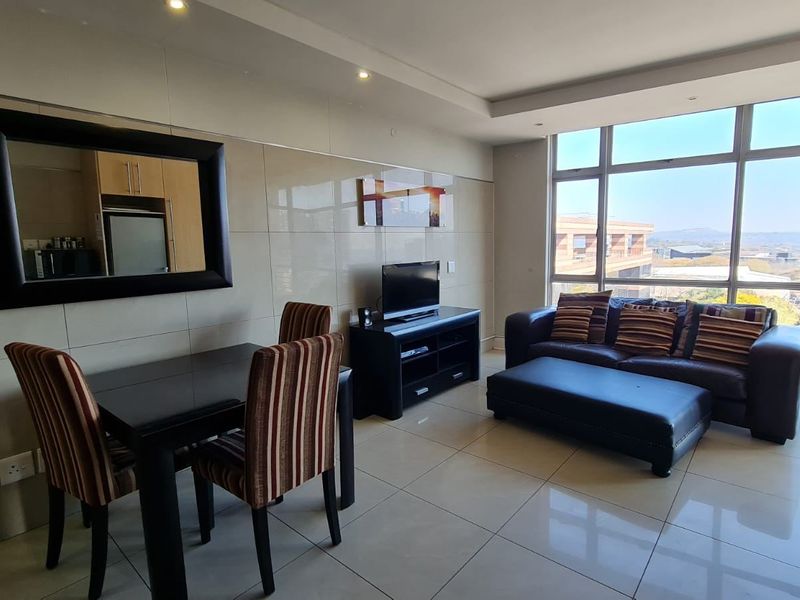 A bright open-plan living and dining area featuring large windows with scenic views, modern tiled flooring, and a sleek tiled feature wall.