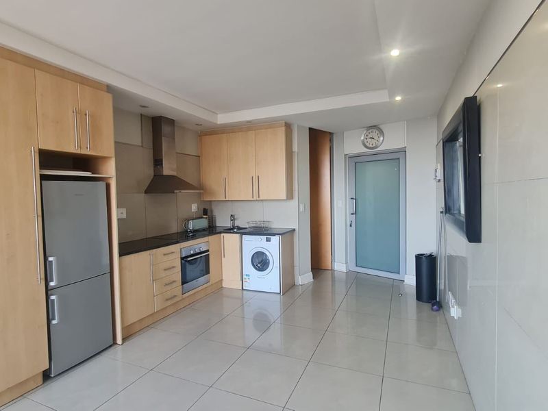 A modern kitchen featuring light wood cabinetry, stainless steel appliances, tiled flooring, and a sleek frosted glass door leading to the rest of the apartment.