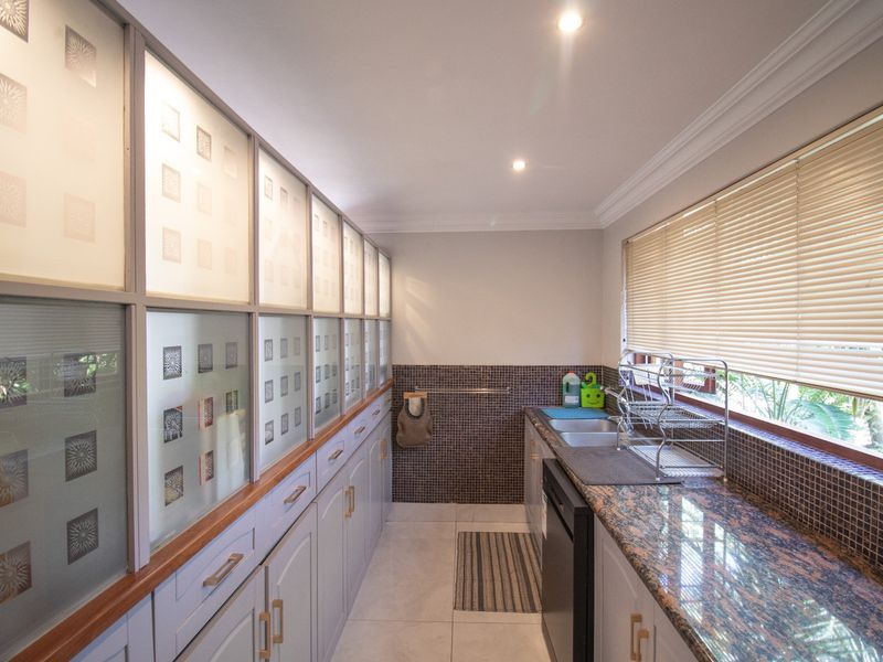 A well-appointed scullery featuring extensive built-in cabinetry with frosted glass detailing, granite countertops, a double sink, and mosaic tile backsplash.