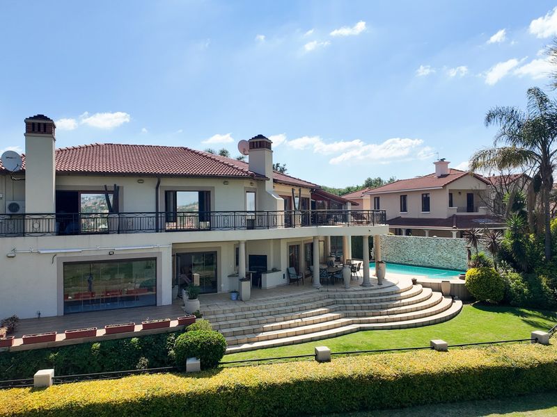Spacious rear elevation featuring a large balcony, covered patio with curved stone steps, swimming pool, and well-maintained landscaped garden.