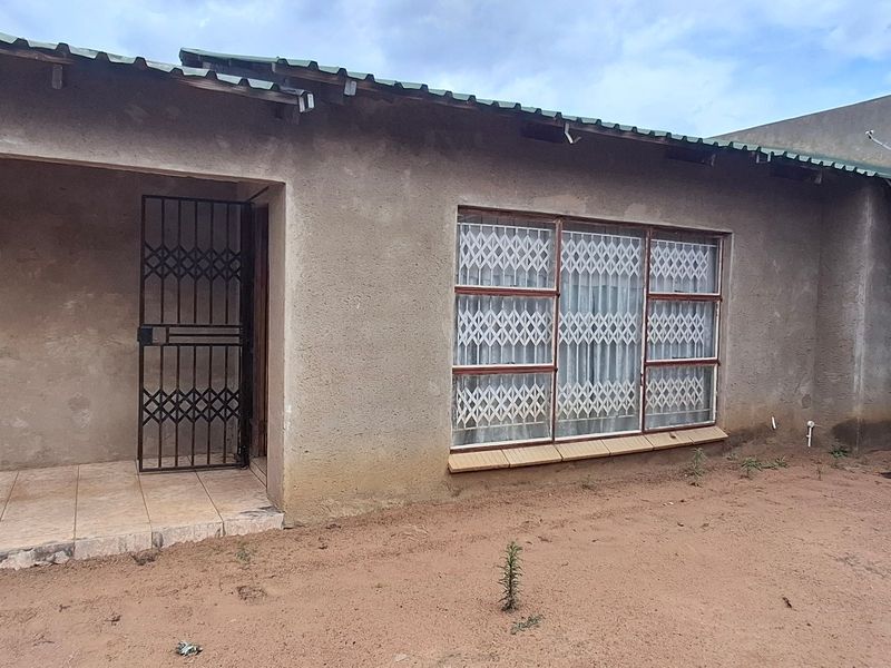 Front elevation of a single-story house featuring a tiled porch, metal security gates, and window bars, with an unfinished plaster exterior finish.