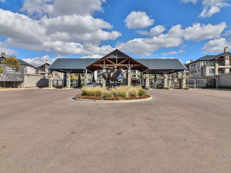 The main entrance to the apartment complex features a secure gatehouse, paved driveway, and a landscaped roundabout with a central sculpture.