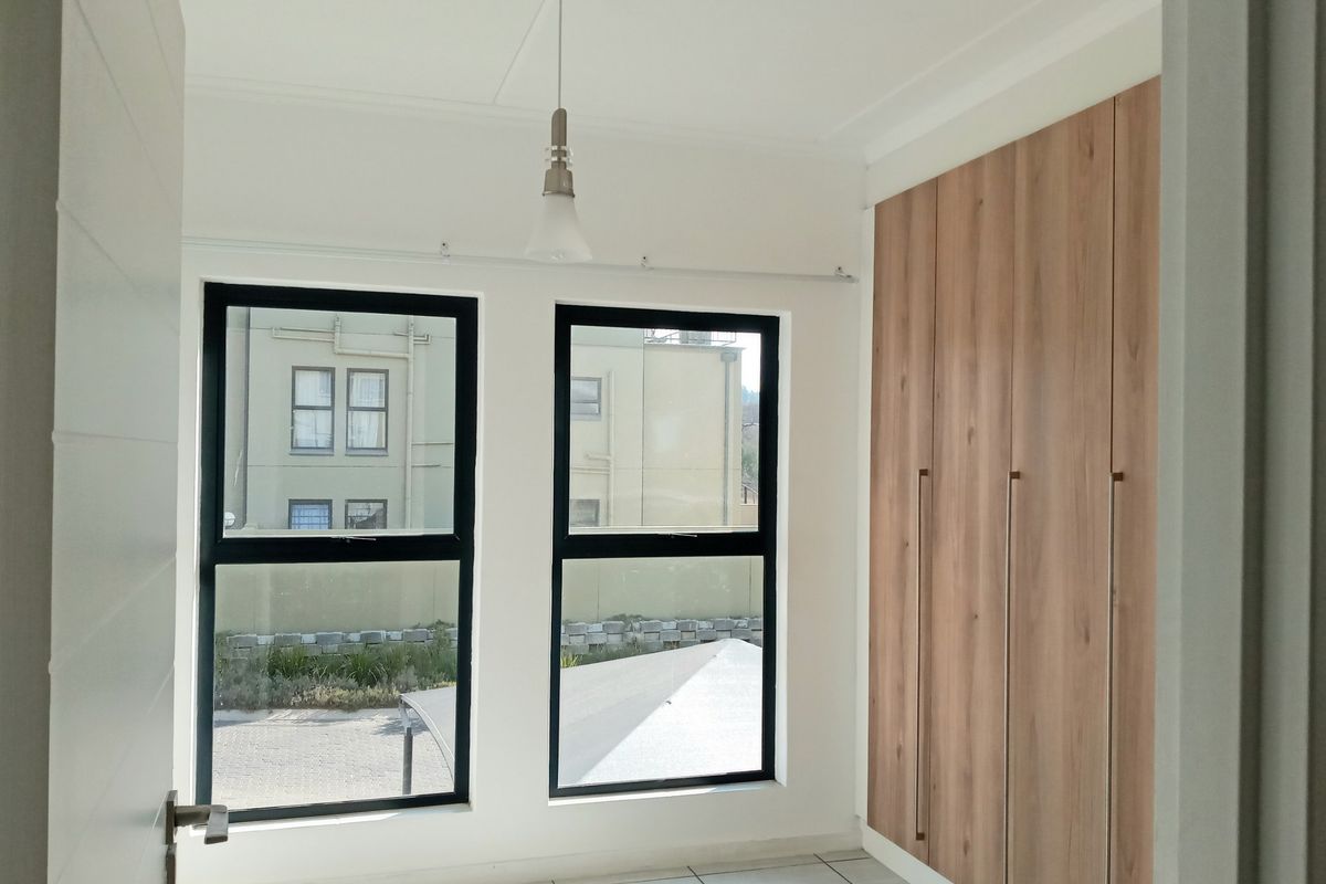 A bright, modern bedroom featuring neutral tiled flooring, large aluminium-framed windows, and sleek, full-height built-in wooden cupboards.