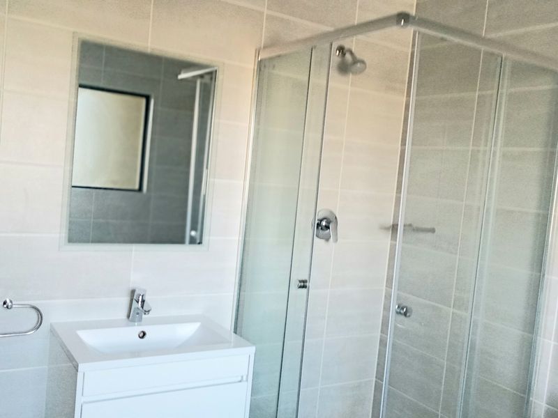 A modern, clean bathroom featuring a white vanity cabinet, a glass-enclosed shower, and neutral wall tiling.
