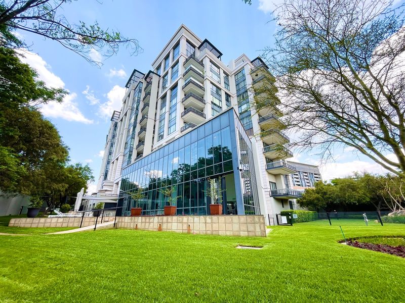 A modern multi-story apartment building featuring a prominent glass facade, private balconies, and well-maintained landscaped grounds.