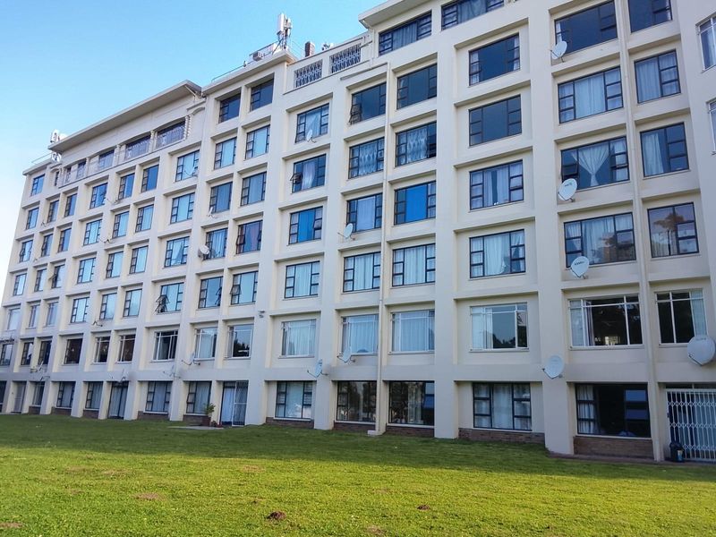 A view of the rear facade of a multi-story apartment building featuring numerous windows and satellite dishes, set against a large communal lawn area.
