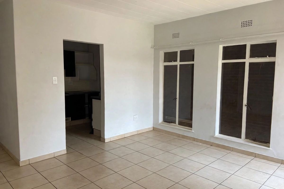 An empty, open-plan living area featuring neutral tiled flooring, large windows for natural light, and direct access to the kitchen area.