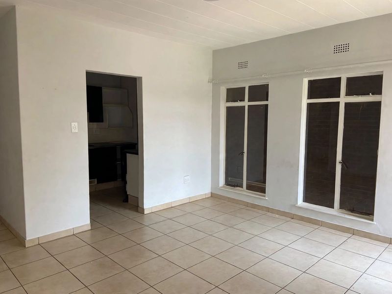 An empty, open-plan living area featuring neutral tiled flooring, large windows for natural light, and direct access to the kitchen area.