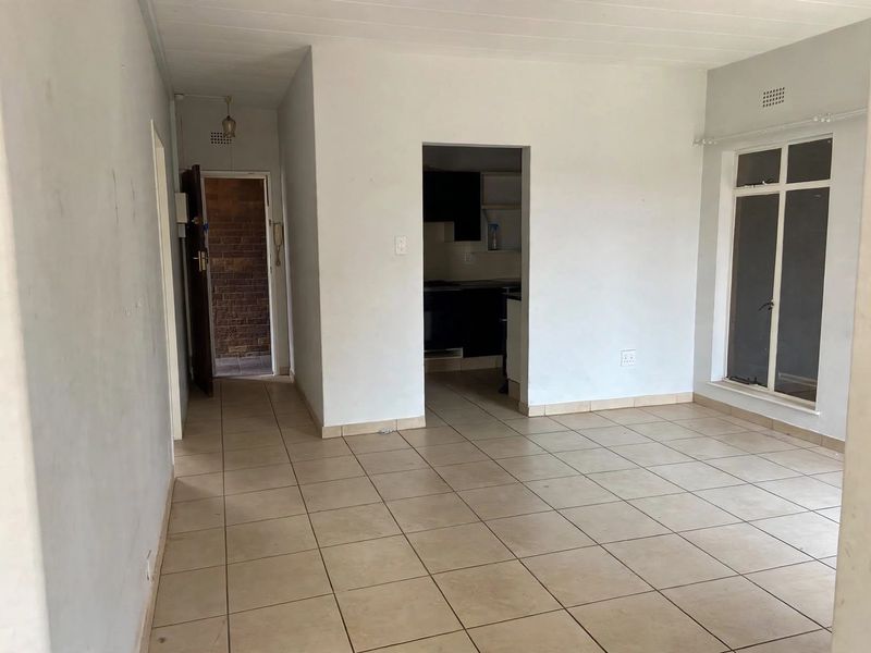 An empty, open-plan living area featuring neutral tiled flooring and direct access to an adjacent kitchen and hallway.