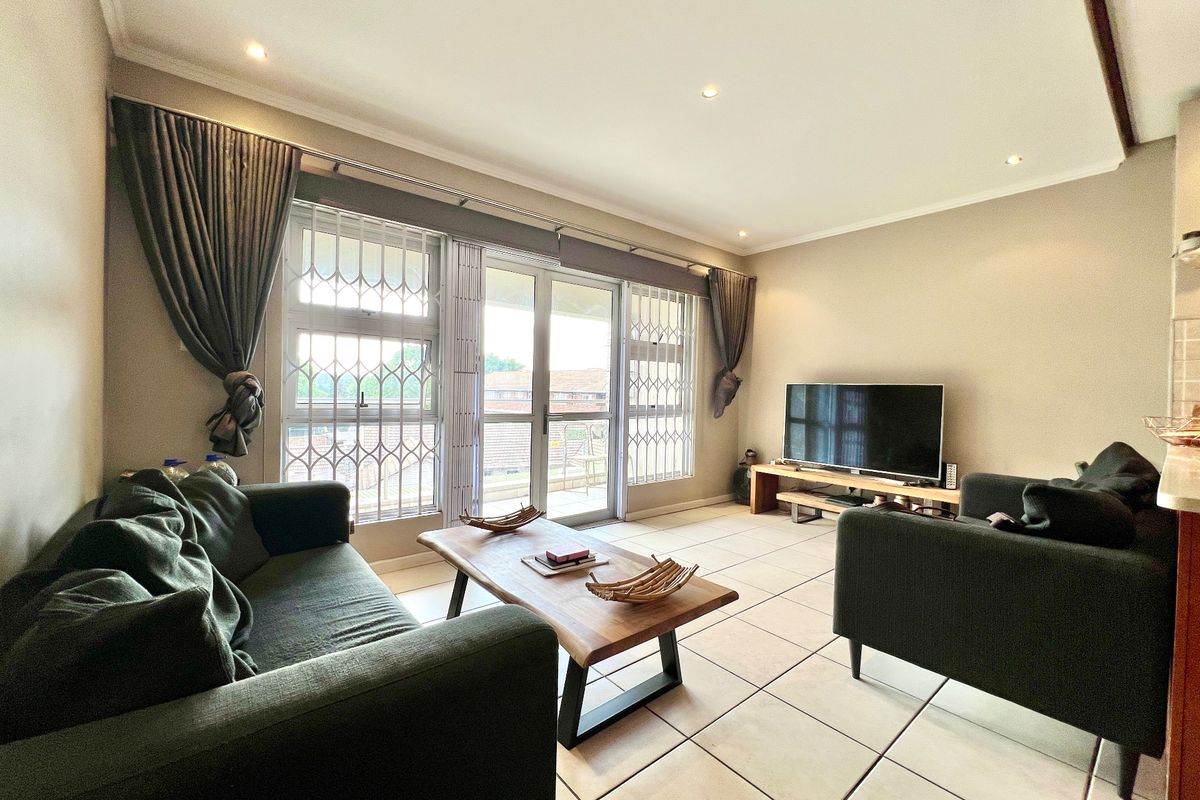 A well-lit living room featuring tiled flooring, recessed downlights, and a sliding glass door providing access to the balcony with security gates.