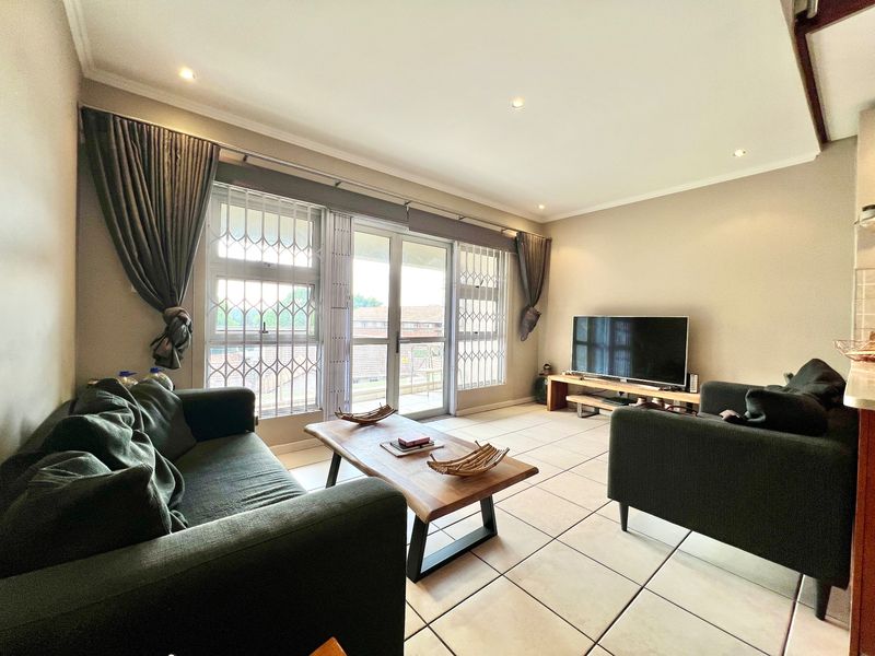 A well-lit living room featuring tiled flooring, recessed downlights, and a sliding glass door providing access to the balcony with security gates.