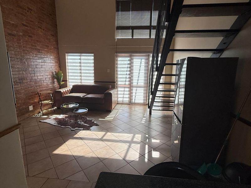 Spacious open-plan living area featuring a striking exposed brick wall, double-volume ceilings, and a modern metal staircase leading to an upper level.