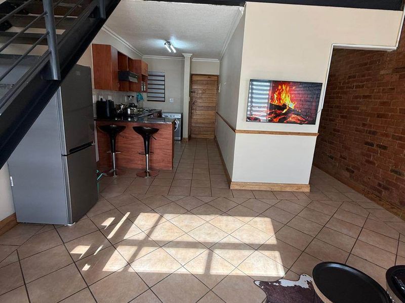 Open plan living area featuring exposed brick walls, tiled flooring, and a metal staircase leading to a mezzanine level, adjacent to a compact kitchen.