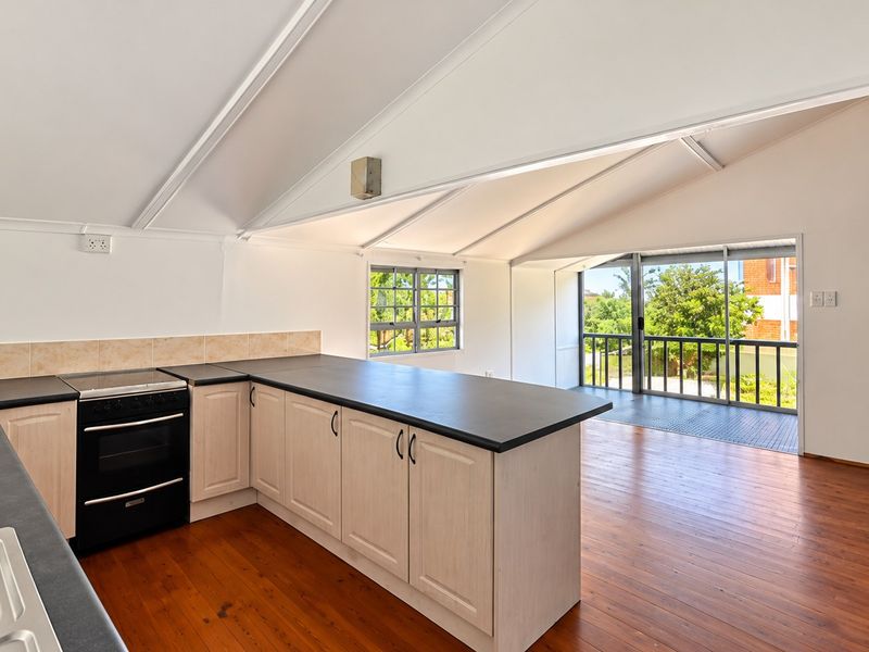 Open plan kitchen and living area featuring timber flooring, vaulted ceilings, and direct access to an outdoor balcony through sliding glass doors.