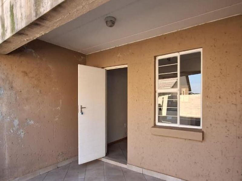 A covered patio area featuring tiled flooring, a security door, and a window with burglar bars, requiring maintenance to the exterior wall finishes.