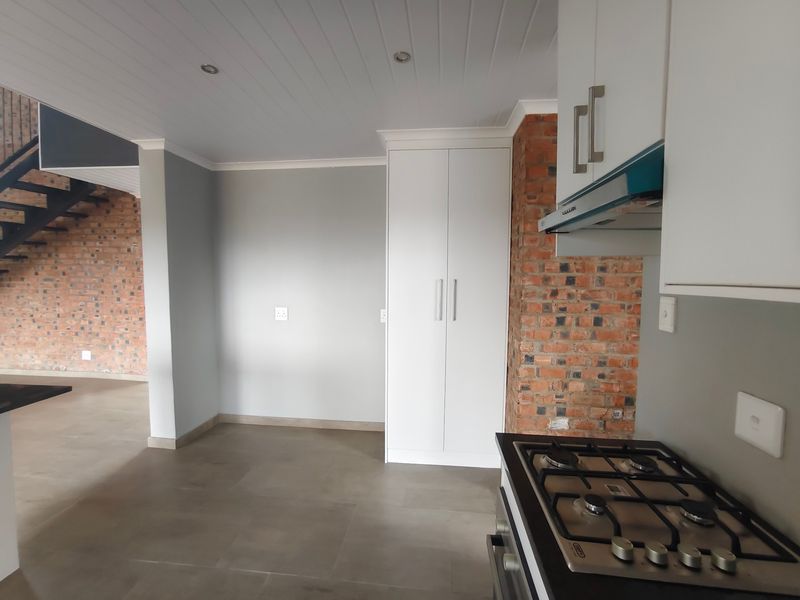 Modern kitchen area featuring exposed brickwork, white cabinetry, a gas hob with extractor, and tiled flooring, seamlessly flowing into an open-plan living space.