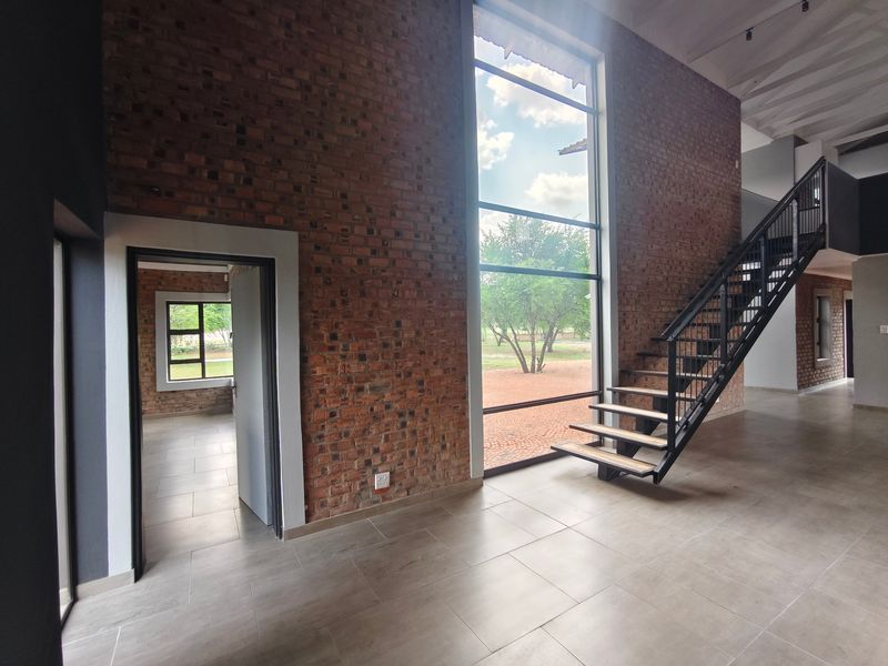 Spacious living area featuring double-volume ceilings, exposed brick walls, large floor-to-ceiling windows, and a modern metal staircase.