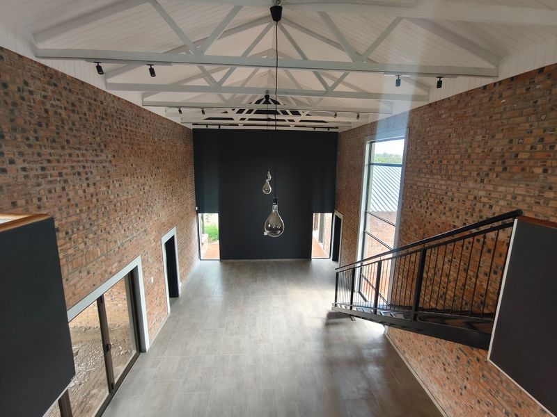 Spacious double-volume living area featuring exposed brick walls, vaulted ceilings with exposed trusses, modern tiled flooring, and a sleek metal staircase.