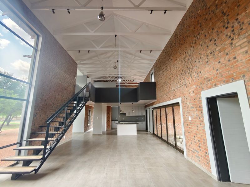 Spacious open-plan living area featuring high ceilings with exposed trusses, industrial-style staircase, mezzanine level, and a modern kitchen with brick wall accents.