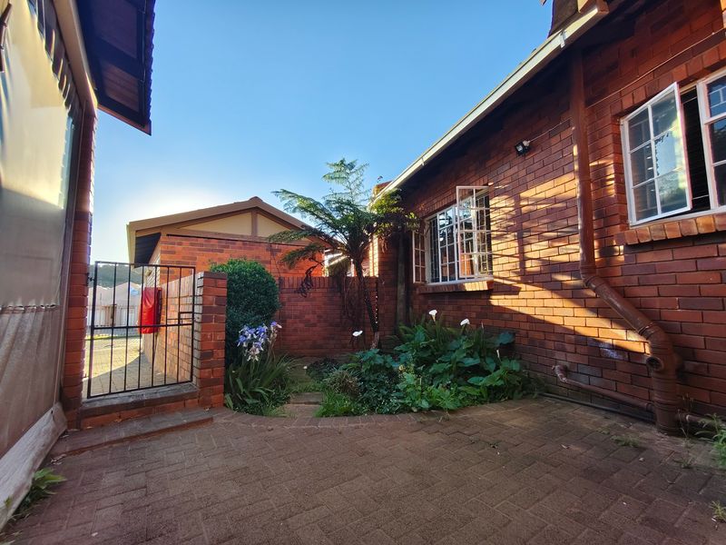 A rear exterior view of a face brick townhouse featuring a paved patio area, established garden beds, and secure metal gate access.