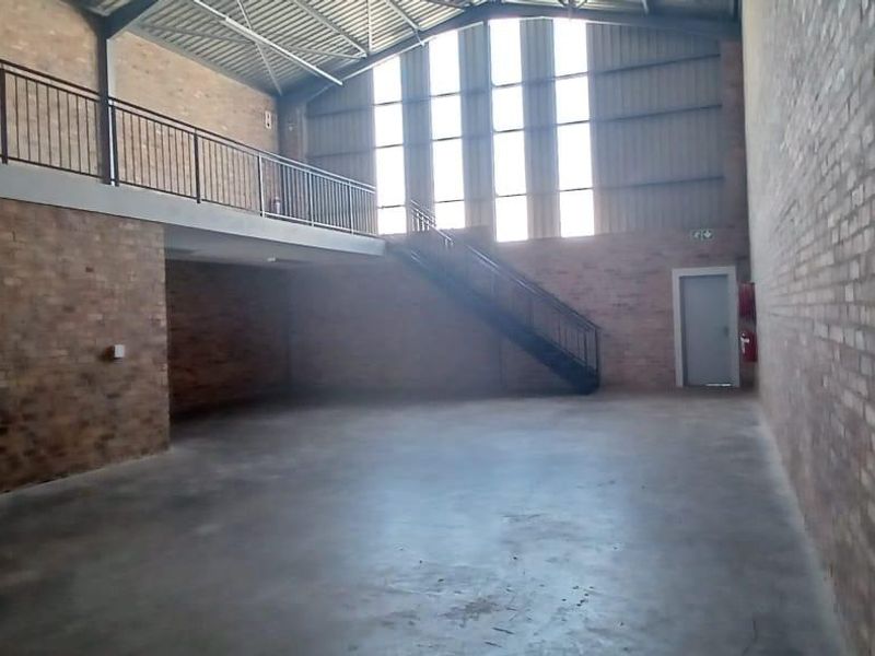 Spacious industrial open-plan area featuring exposed brick walls, concrete flooring, a mezzanine level with metal staircase, and high-level windows providing ample natural light.
