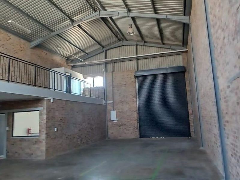 Spacious industrial warehouse unit featuring exposed brick walls, a mezzanine level, high ceilings with steel trusses, and a large roller shutter door.