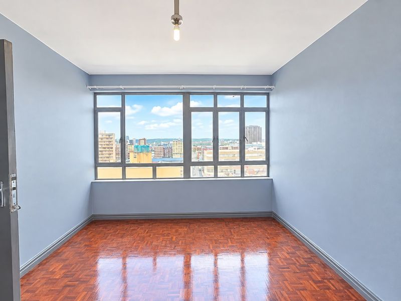 An empty bedroom featuring polished parquet flooring, blue-painted walls, and a large window offering an expansive city view.
