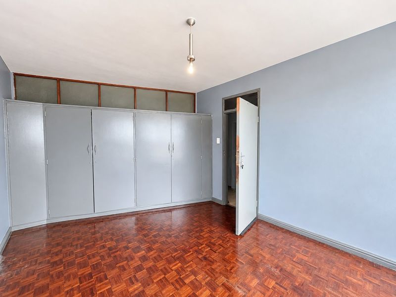 A bright, empty bedroom featuring classic parquet flooring, extensive built-in cupboards with overhead storage, and a neutral blue wall finish.