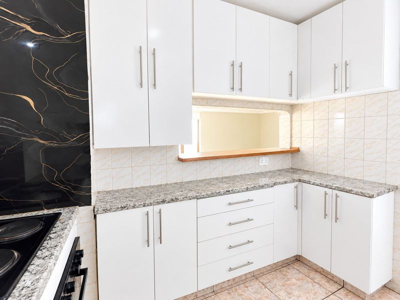 A clean, functional kitchen featuring white cabinetry, granite countertops, a ceramic tile backsplash, and a convenient pass-through window to the adjacent living area.