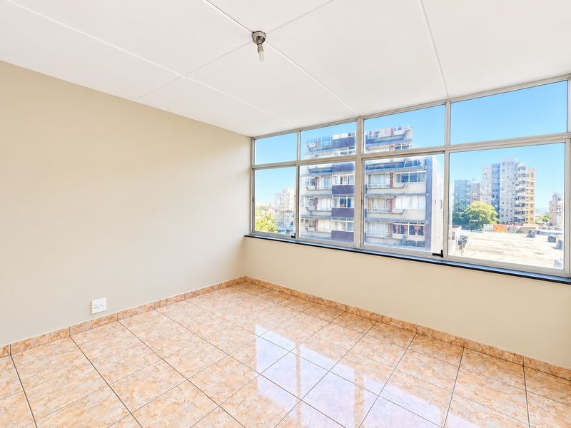 A bright, empty living area featuring large windows with city views, tiled flooring, and a drop ceiling, offering a blank canvas for personalization.