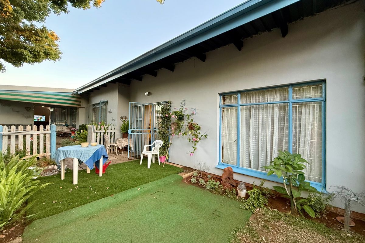 Rear exterior view of the house featuring a paved patio area, large windows with blue frames, and a secure metal entrance gate.