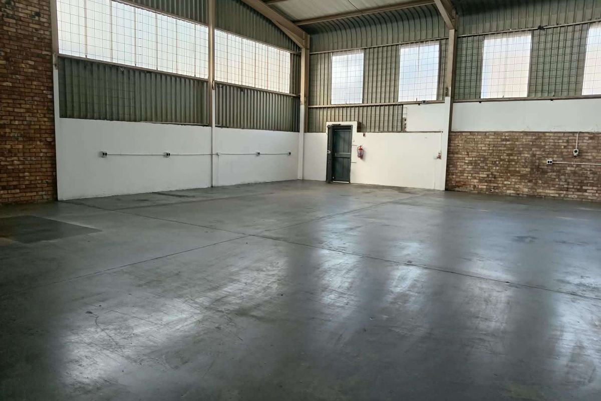 Spacious industrial warehouse unit featuring exposed brick walls, high-clearance corrugated metal ceiling, concrete flooring, and large industrial windows providing ample natural light.