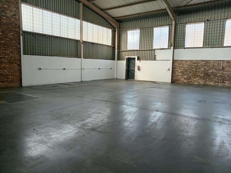 Spacious industrial warehouse unit featuring exposed brick walls, high-clearance corrugated metal ceiling, concrete flooring, and large industrial windows providing ample natural light.