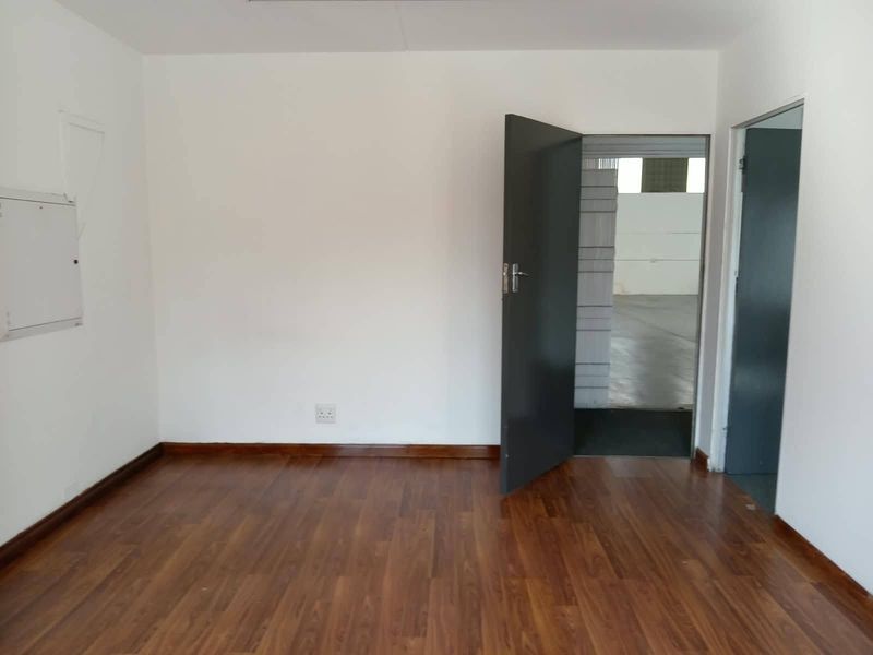 A clean, empty office space featuring laminate wood flooring and direct access to an adjacent industrial area.
