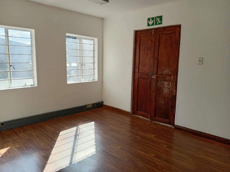 A vacant office space featuring laminate flooring, security-barred windows, and wooden double doors with emergency exit signage.