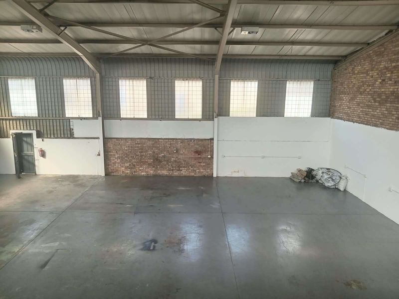 Spacious industrial warehouse unit featuring high ceilings with exposed steel trusses, concrete flooring, and a combination of brick and painted wall finishes.