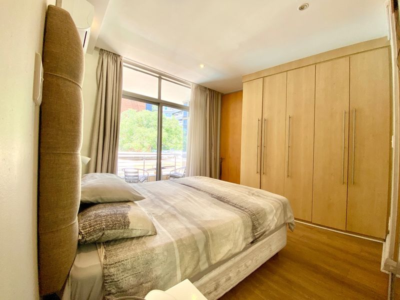 A well-maintained bedroom featuring light wood-toned built-in wardrobes, laminate flooring, and a sliding glass door providing access to a balcony.