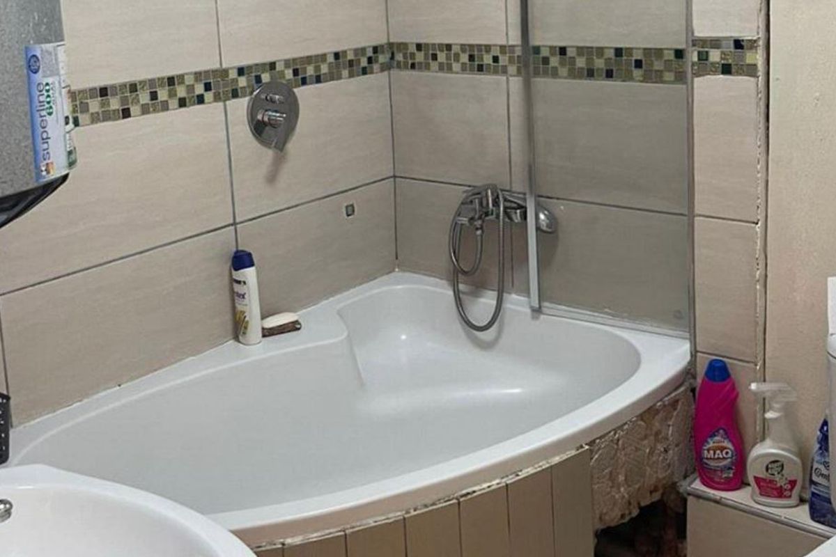 A compact bathroom featuring a built-in bathtub with shower mixer, toilet, and basin, requiring maintenance to the bathtub apron and tiling.