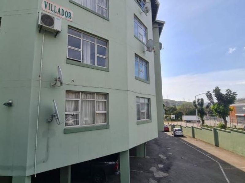 Exterior view of the Villador apartment complex featuring a multi-story facade, paved driveway, and boundary wall with nearby commercial amenities.
