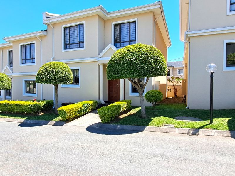 A well-maintained townhouse exterior featuring a clean plastered facade, manicured garden, and paved entrance walkway within a secure residential complex.