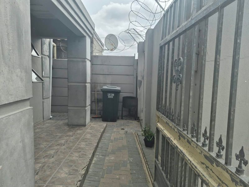 A side entrance area featuring a metal security gate, paved walkway, tiled porch, and perimeter walling secured with razor wire.