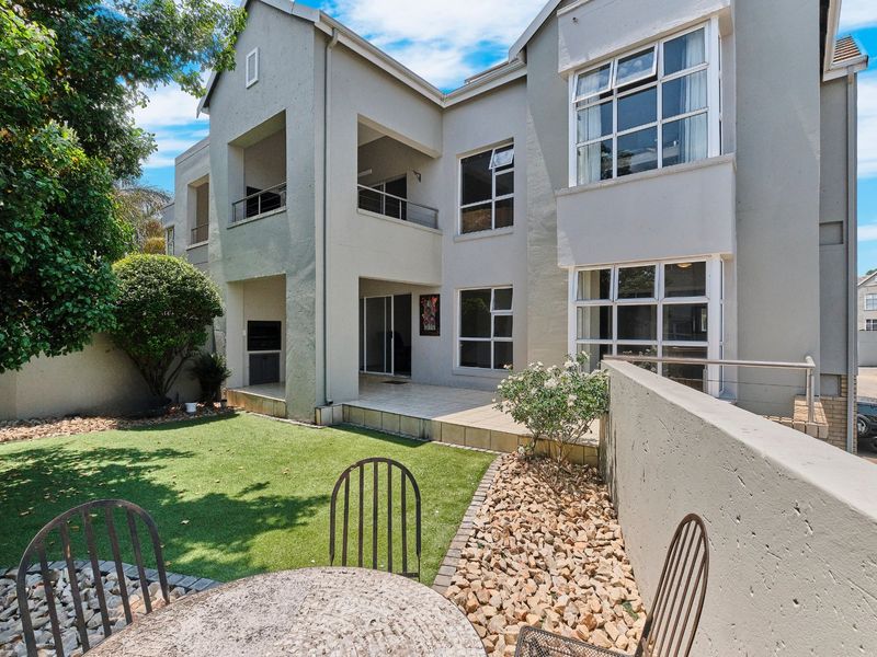 Rear view of the apartment building featuring a tiled patio, built-in braai area, and a private garden with artificial lawn.