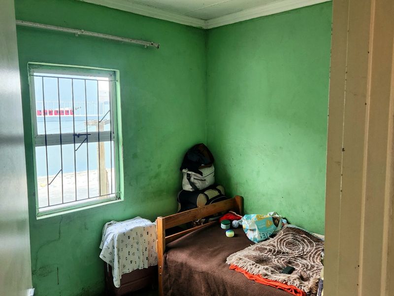 A compact bedroom featuring green walls, a window with security bars, and an unfinished concrete floor, requiring cosmetic updates and refurbishment.