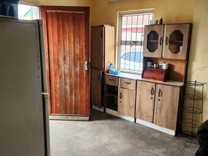 A dated kitchen area featuring basic built-in cabinetry, a timber door, and security bars on the window, requiring cosmetic updates and floor finishing.