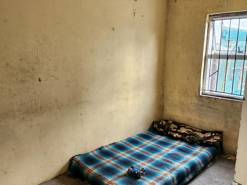 A small bedroom featuring concrete flooring and security bars on the window, currently requiring significant wall repairs and cosmetic refurbishment.