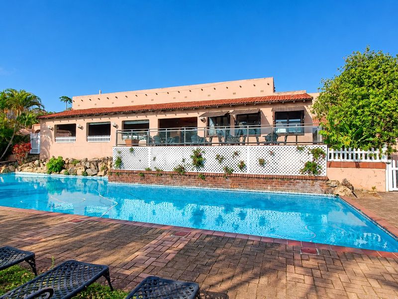 A well-maintained outdoor pool area featuring a large swimming pool, brick paved surround, and a raised patio with a glass balustrade.